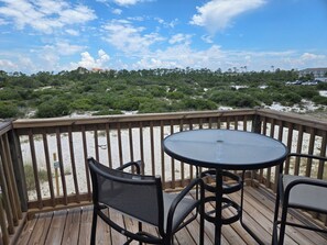 Una terraza de madera con una mesa redonda y dos sillas, con vista a una playa de arena y vegetación escasa.