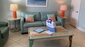 Una sala de estar con un sofá, dos mesas auxiliares con lámparas, una mesa de centro con un libro y una bolsa, y un cuadro en la pared.