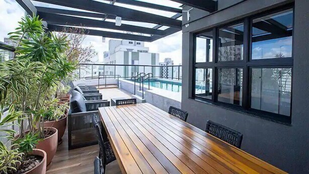 Terraço no telhado com mesa e cadeiras de madeira, plantas em vasos e vista para a cidade.