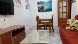 A living room with a glass-top coffee table, wooden furniture, a TV mounted on the wall, and a painting of a coastal scene. - Apartamento Candari Playa (Cádiz)