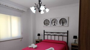 A bedroom with a bed, two bedside tables, a chandelier, and three wall-mounted tree artwork. - Apartamento Candari Playa (Cádiz)