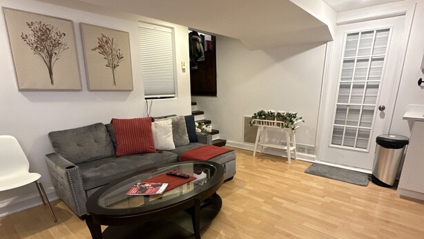 A modern living room with a grey sofa, a glass coffee table, and wooden flooring. There are two framed floral artworks on the wall, a white door, and a small white table with a plant.