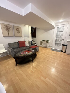 Een moderne woonkamer met een grijze bank, een glazen salontafel en houten vloeren. Twee ingelijste bloemwerken hangen aan de muur, een witte deur en een klein witte tafeltje met een plant.