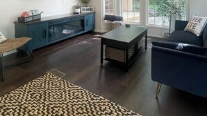A modern living room with a dark wood floor, a patterned rug, a blue sofa, a coffee table, and a TV mounted on the wall. - River's Edge Tiny Home | Expansive Deck (Park Falls)