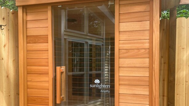 A wooden sauna with a sliding glass door, two white lanterns, and the text "sunlighters Amplify" on the glass. - River's Edge Tiny Home | Expansive Deck (Park Falls)
