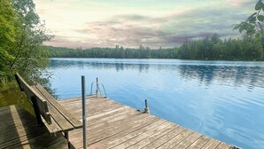 A wooden dock extending over calm water with a forested shoreline in the background. - River's Edge Tiny Home | Expansive Deck (Park Falls)