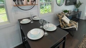A dining area with a dark wooden table set for four, a hanging pendant light, and a view of greenery outside. - River's Edge Tiny Home | Expansive Deck (Park Falls)