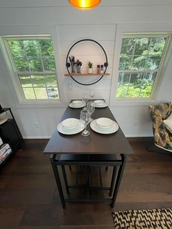 Un área de comedor con una mesa dispuesta para dos, un estante sobre la mesa y vistas a la vegetación a través de las ventanas.