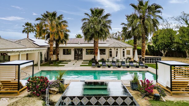 A pool area with lounge chairs, palm trees, and a house in the background.