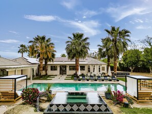 Zona de piscina con sillas de descanso, palmeras y una casa al fondo.