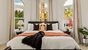 A bedroom with a large bed, two bedside tables with lamps, and a view of trees and flowers through arched windows.