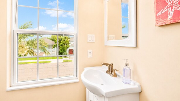 A bathroom with a white sink, a mirror, and a window with a view of a house and palm trees. - Heated Pool | Pet Friendly | Fire Pit | Near Beach (Galveston)