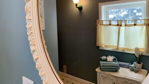 A bathroom with a dark wall, a white vanity with a mirror, a window with a yellow curtain, and a wall-mounted light fixture. - In-Town Home, Hot Tub, Wifi & Minutes to Skiing (Red Lodge)