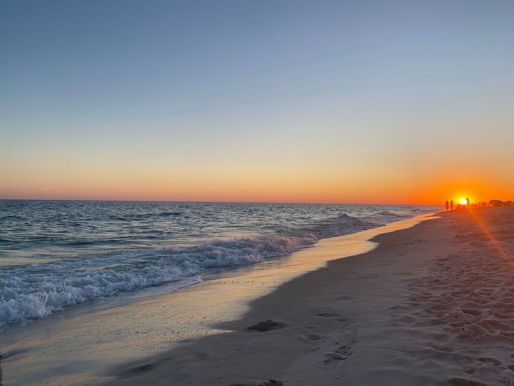 Een strand bij zonsondergang met golven, voetafdrukken en mensen in de verte.