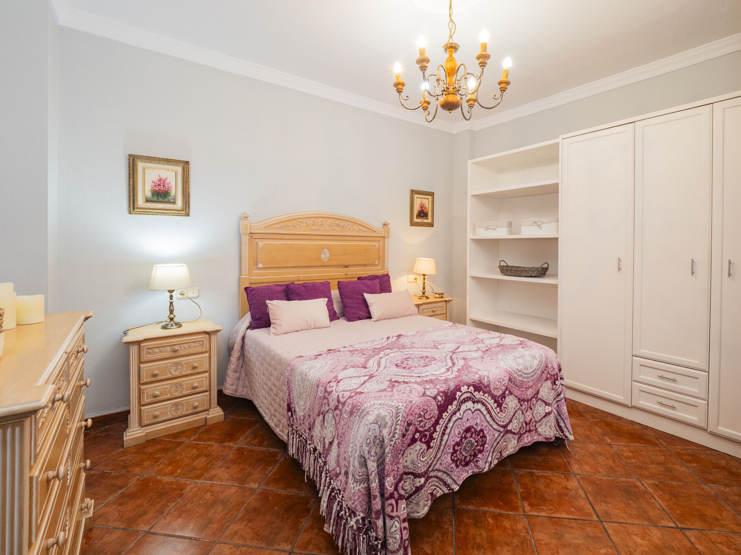 Ein Schlafzimmer mit einem hölzernen Bett, zwei Nachttischen mit Lampen, einer Kandelaberleuchte, einem Kleiderschrank und einem Regal mit Aufbewahrungsboxen.