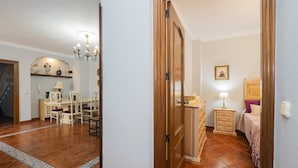 Una habitación con puerta de madera, cama con cabecera, mesita de noche con lámpara y cómoda.