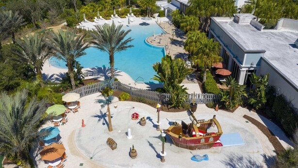 An aerial view of a resort with a large swimming pool, palm trees, and a play area with a pirate ship. - Charming 3Bd Close To Disney At Enclaves At Festival 745 (Davenport)