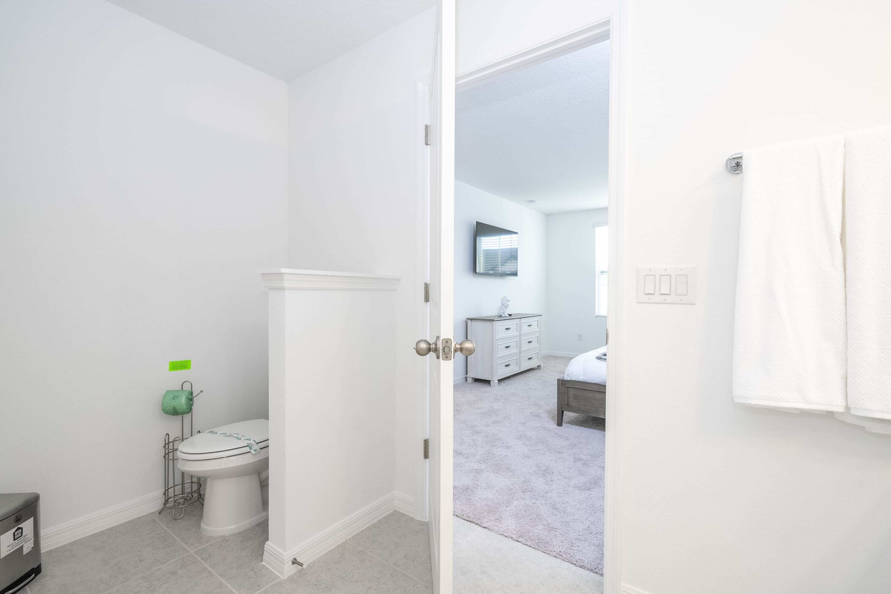 A bathroom with a toilet, a sink, and a shower area. There is a door leading to a bedroom with a bed and a dresser.