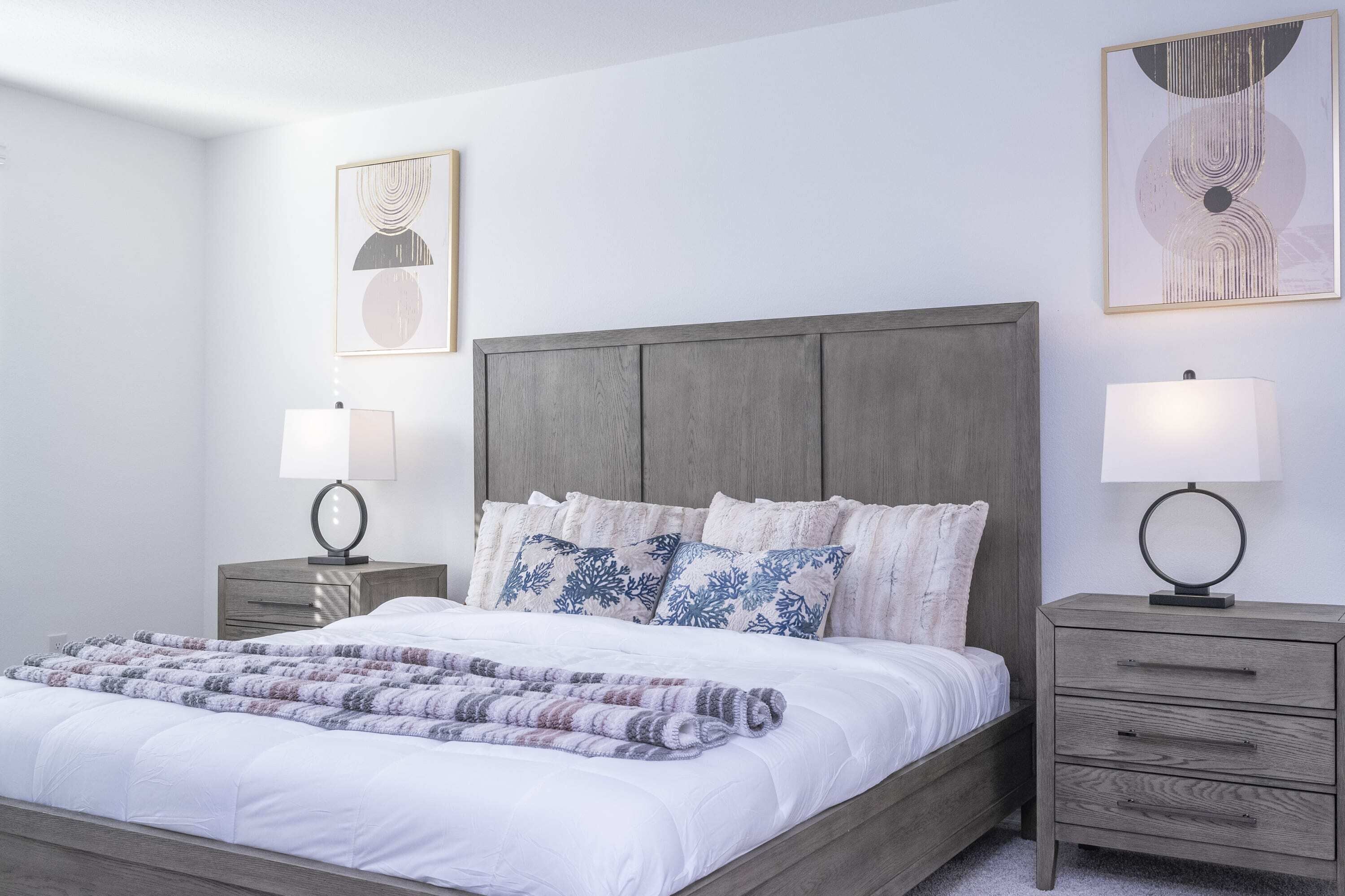 Una cama bien hecha con edredón blanco y manta decorativa, flanqueada por dos mesitas de noche con lámparas, frente a una pared lisa con dos cuadros.
