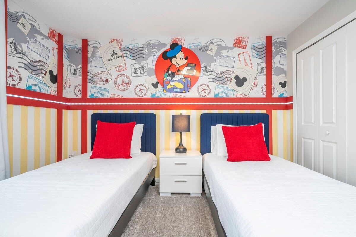 A hotel room with two beds, each with red pillows, a nightstand with a lamp, and a wall decorated with travel-themed artwork.