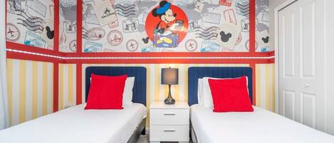 A hotel room with two beds, each with red pillows, a nightstand with a lamp, and a wall decorated with travel-themed artwork.