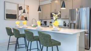 A modern kitchen with a central island, bar stools, pendant lighting, stainless steel appliances, and artwork on the wall. - Charming 4Bd W/ Private Pool @ Storey Lake Resort 2653 (Kissimmee)