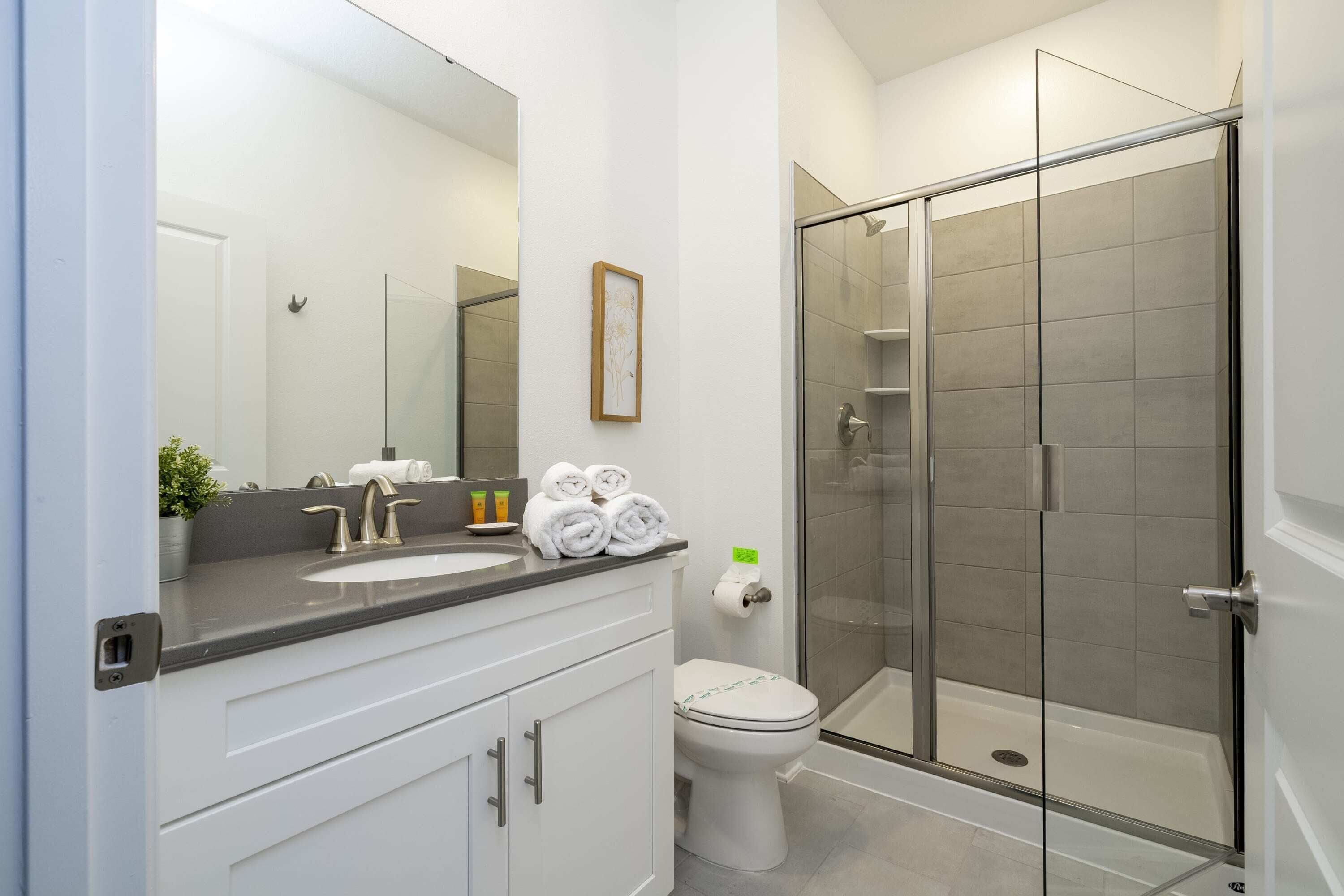 A modern bathroom with a glass-enclosed shower, a white sink with a dark countertop, and a toilet with a roll of toilet paper.
