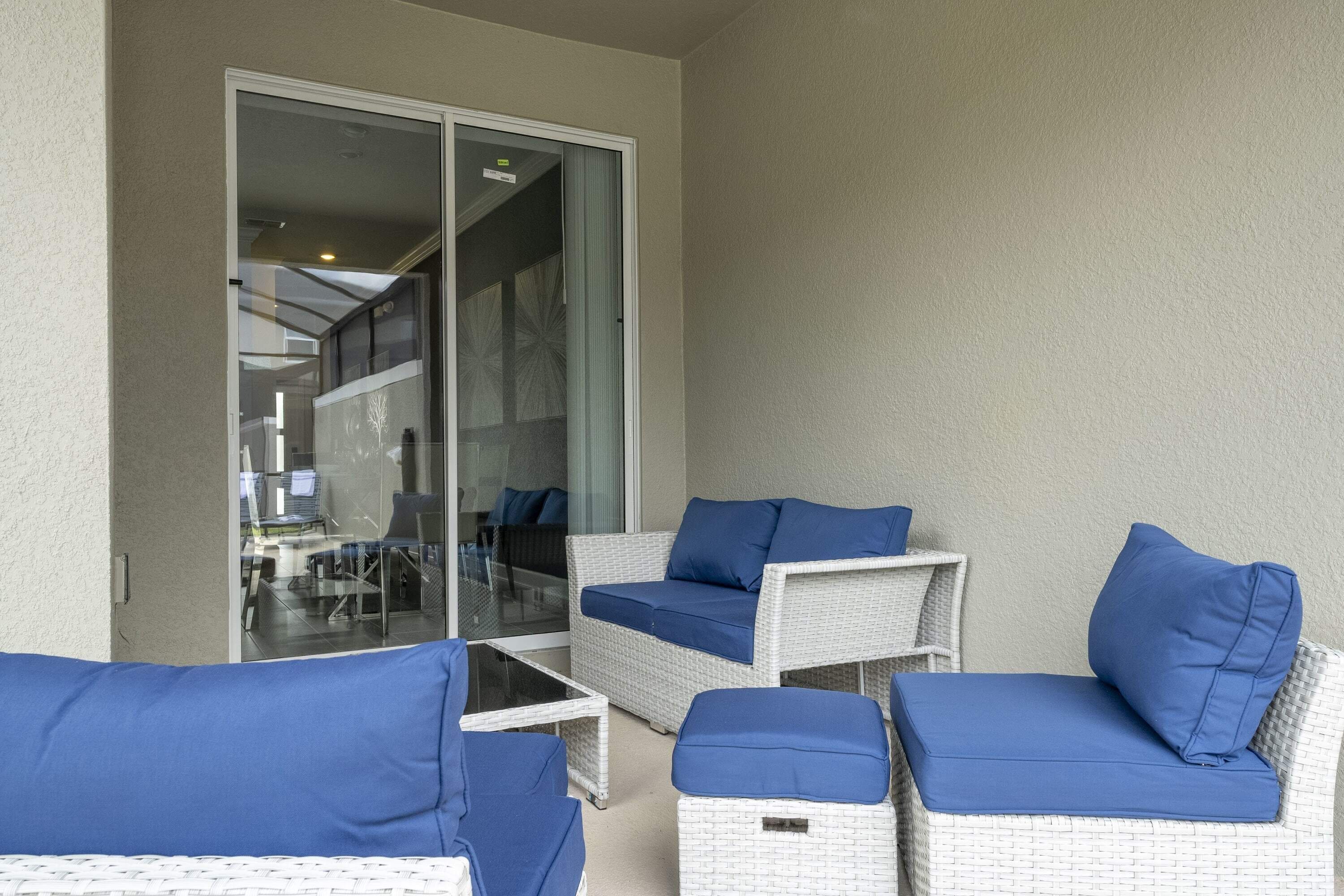 Een balkon met rieten meubilair en blauwe kussens, een glazen deur die leidt naar een binnenruimte, en een witte, gestructureerde muur.
