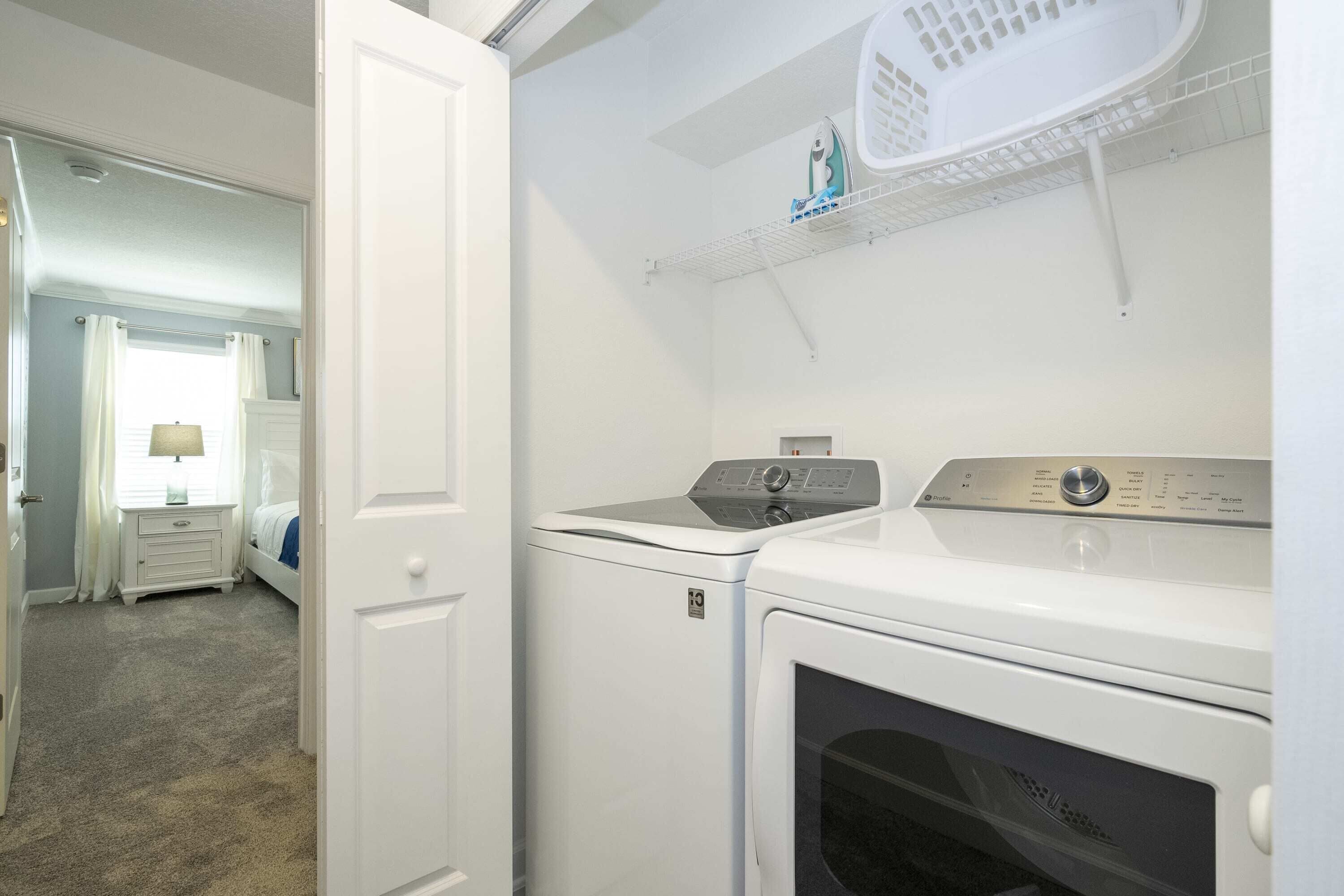 A laundry room with a front-loading washer and dryer, a shelf with laundry baskets, and a door leading to a bedroom.