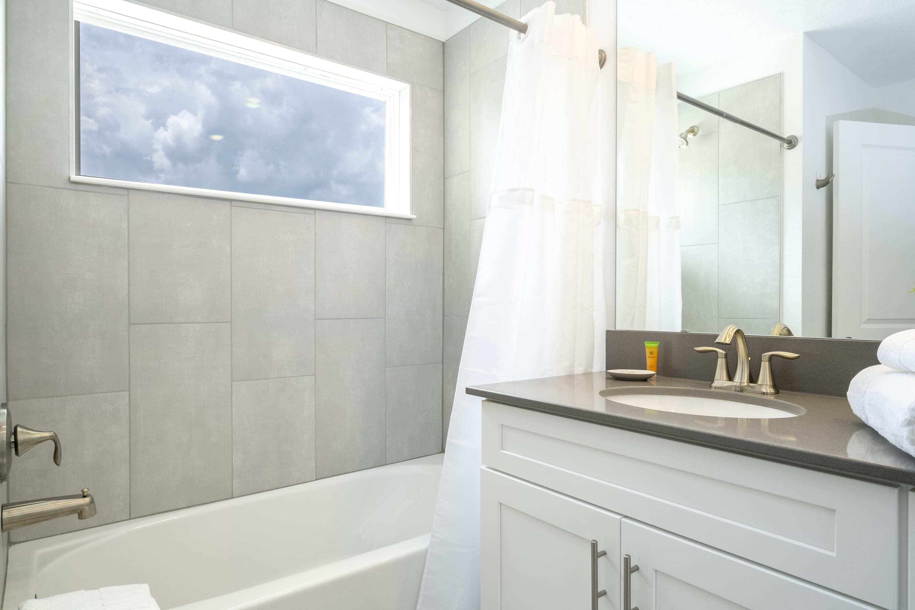 A modern bathroom with a bathtub, a window with a view of the sky, a sink with a mirror, and a shower curtain.