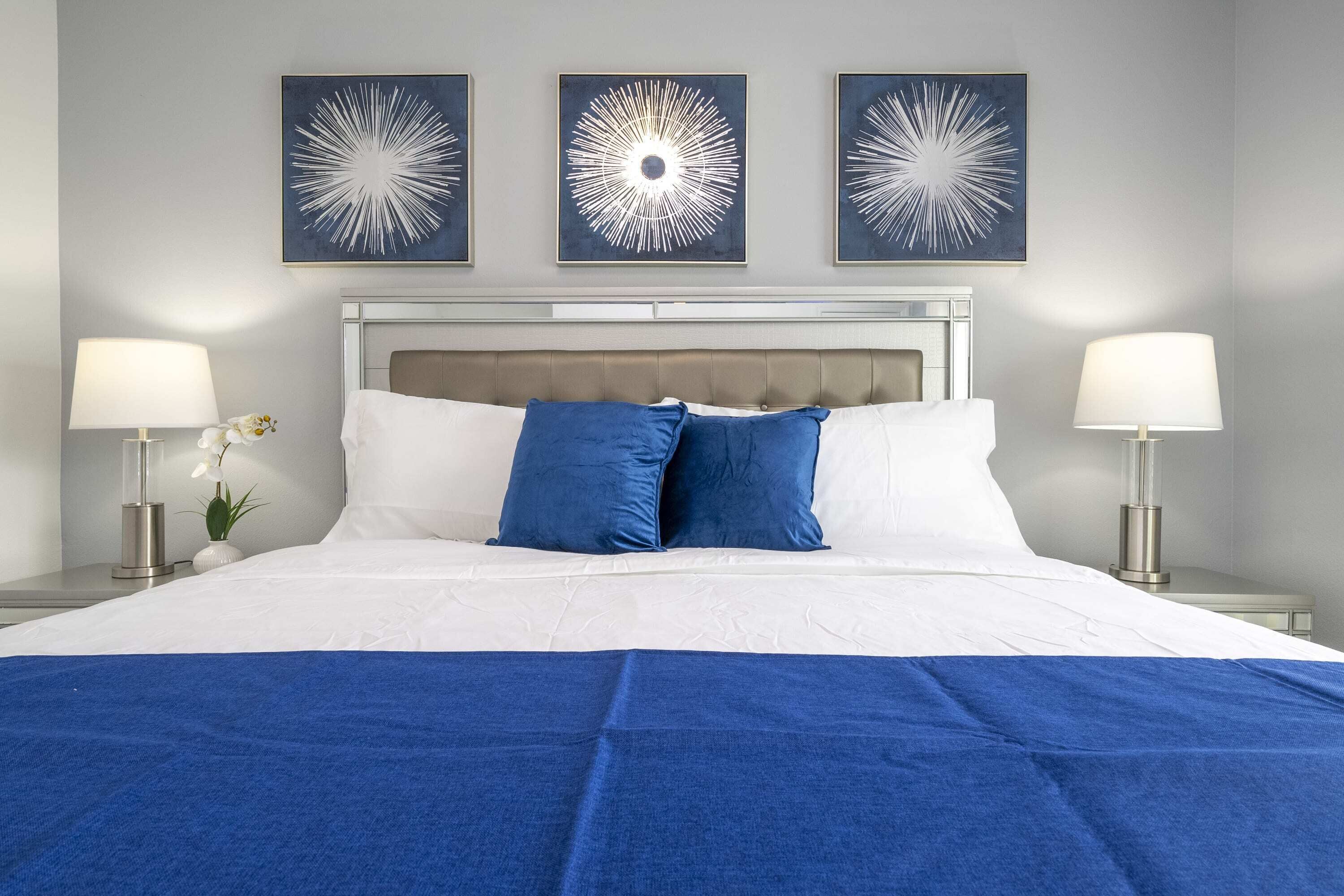 Ein ordentlich bezogenes Bett mit weißer und blauer Bettwäsche, das von zwei Nachttischen mit Lampen und einer Blumenvase eingerahmt wird.