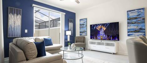 A modern living room with a beige sofa, a glass coffee table, a ceiling fan, and a flat-screen TV.