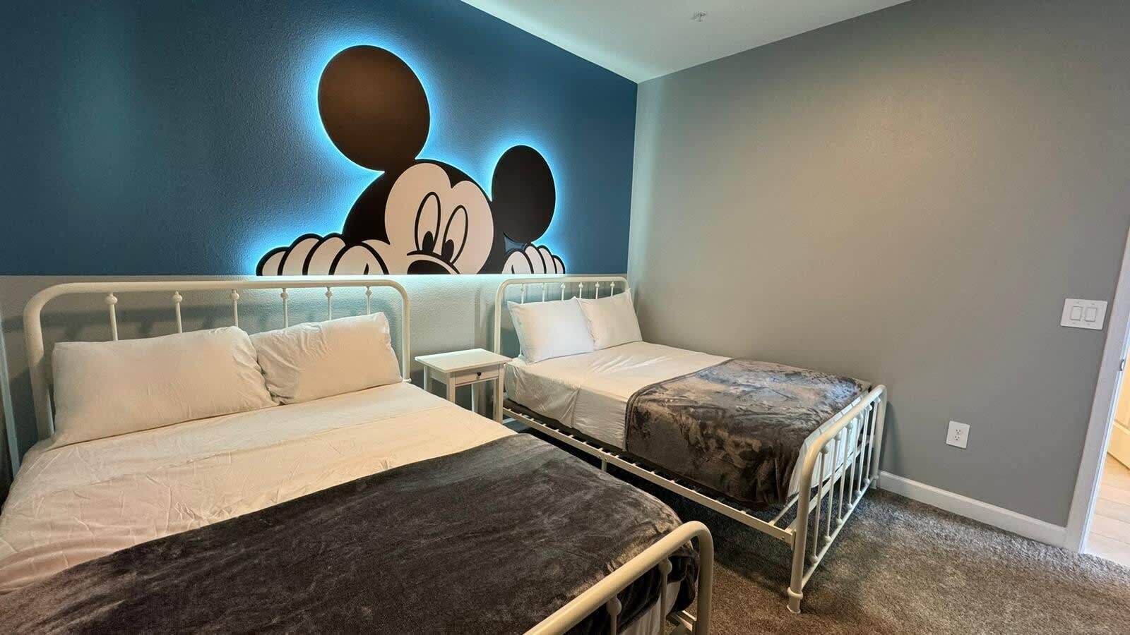 A room with two beds, a Mickey Mouse mural on the wall, and a small bedside table.