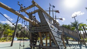 A wooden play structure with ropes and a slide, surrounded by palm trees and a clear sky.