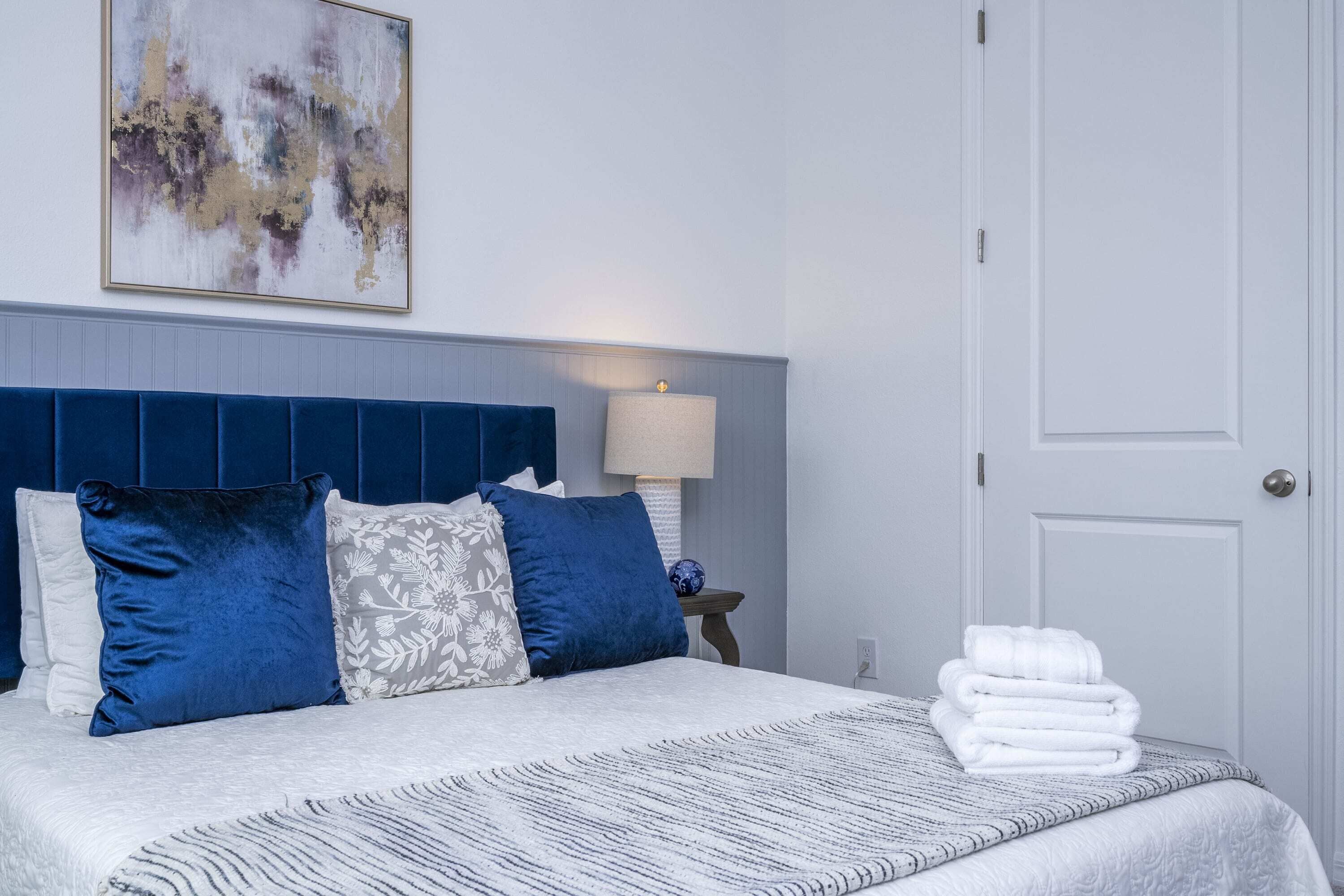 A neatly made bed with a blue headboard, white and patterned pillows, and a stack of white towels on the side.