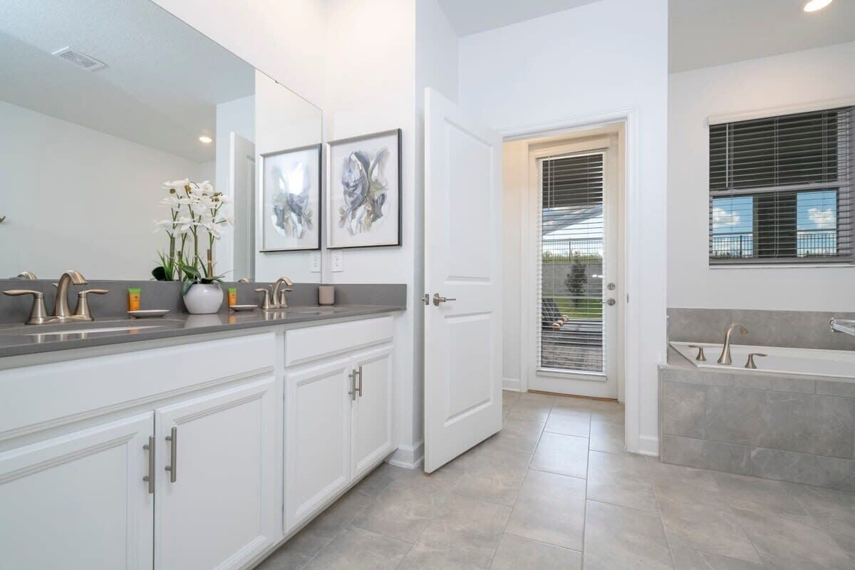 A modern bathroom with a large mirror, white cabinetry, a bathtub, and a window with blinds.