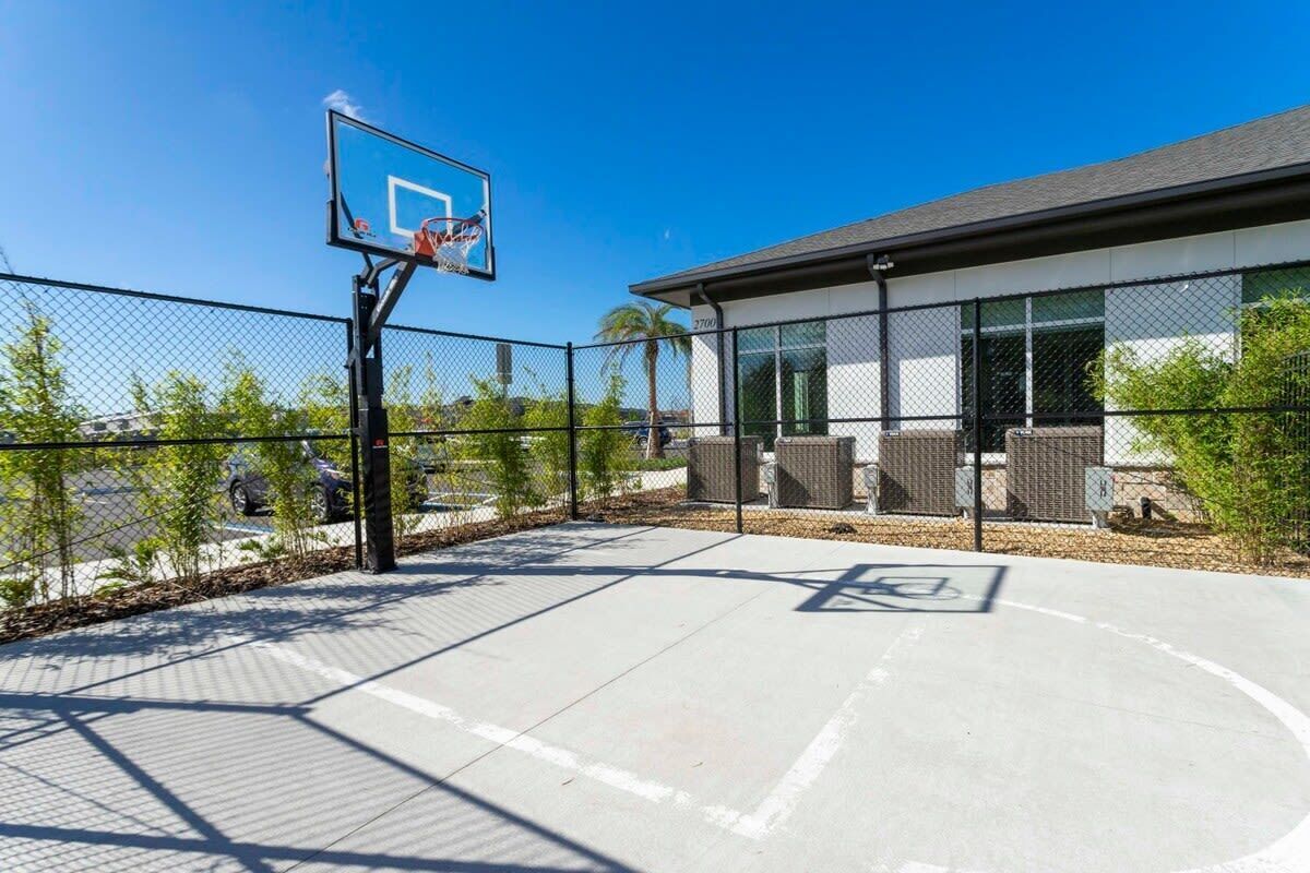 Een basketbalveld buiten met een ring, omgeven door een hek van gaas, en een gebouw met grote ramen op de achtergrond.