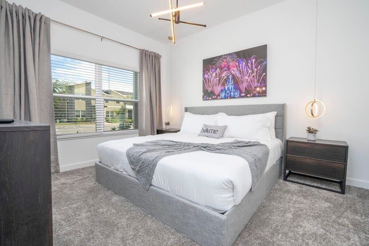 A modern bedroom with a large bed, a nightstand, a wall-mounted light, and a window with curtains.