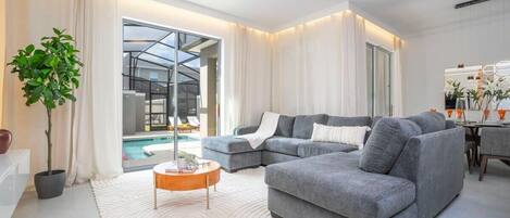 Una sala de estar moderna con un sofá seccional gris, una mesa de centro redonda y un ventanal con vistas a una piscina.