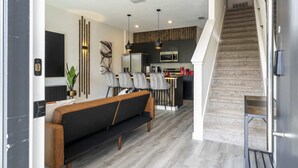 A modern living room with a sofa, a dining area with chairs, a kitchen with a refrigerator, and a staircase.