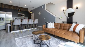 A modern kitchen with a stainless steel refrigerator, microwave, and sink. There is a dining area with a bar stool and a round table. A brown leather sofa is in the living area with a large mirror and wall sconces.