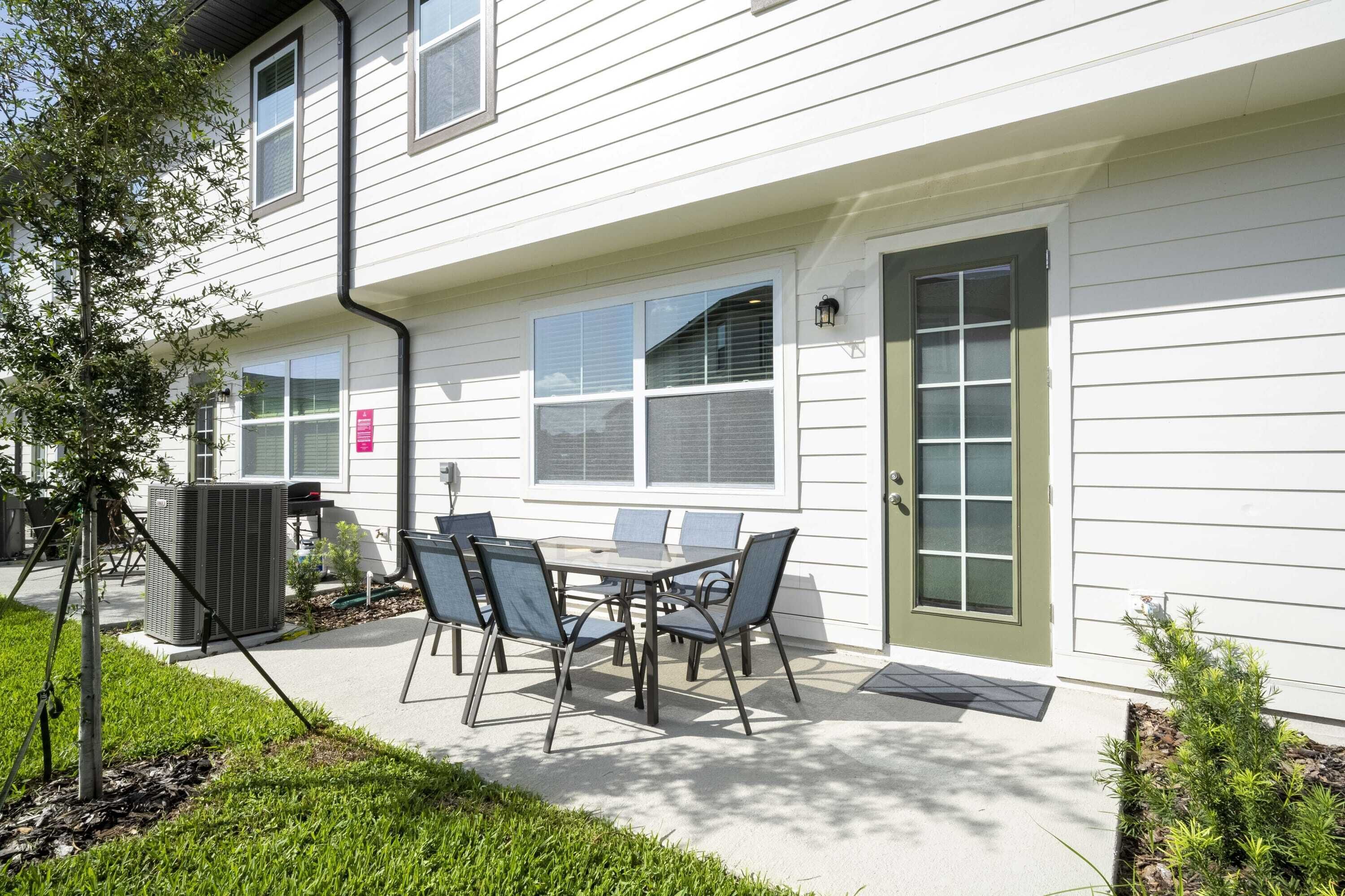 A patio with a table and chairs, a small tree, and a building with white siding and a green door.