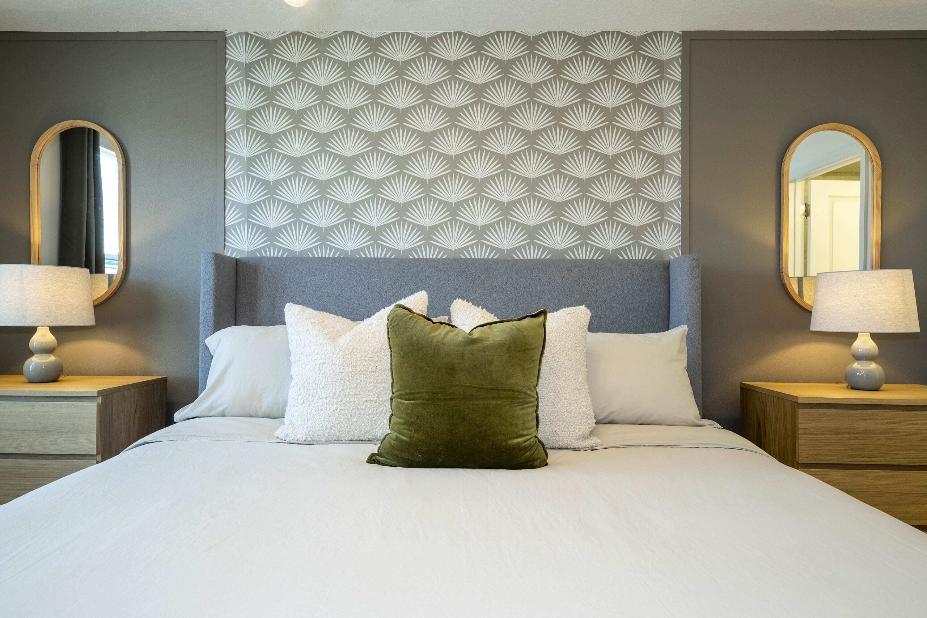A neatly made bed with white linens and a green accent pillow, flanked by two bedside tables with lamps and a patterned headboard.