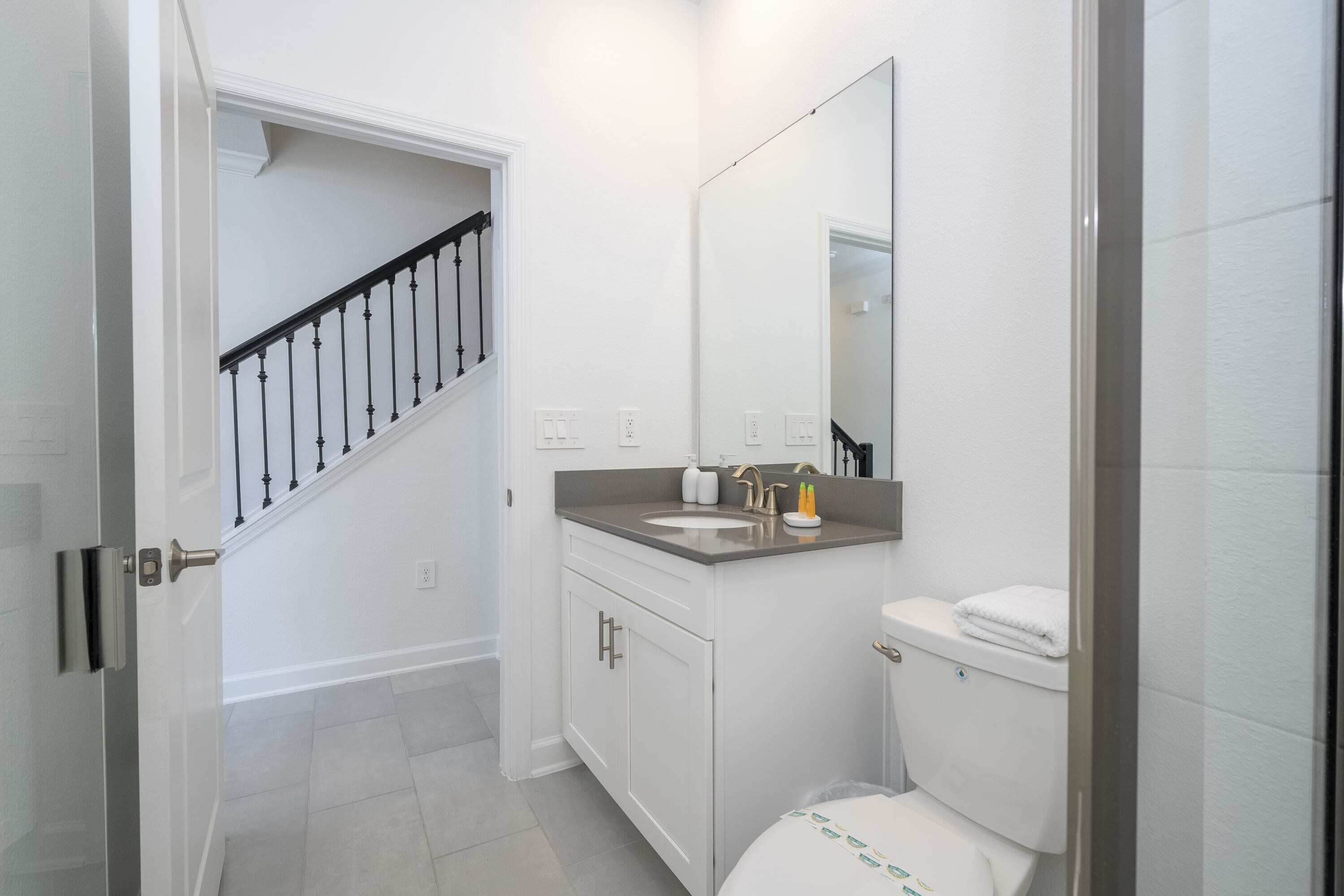 Un baño con inodoro blanco, lavamanos y espejo sobre él. Se ve una escalera que conduce a otra habitación.