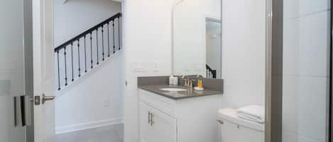 Un baño con inodoro blanco, lavamanos y espejo sobre él. Se ve una escalera que conduce a otra habitación.