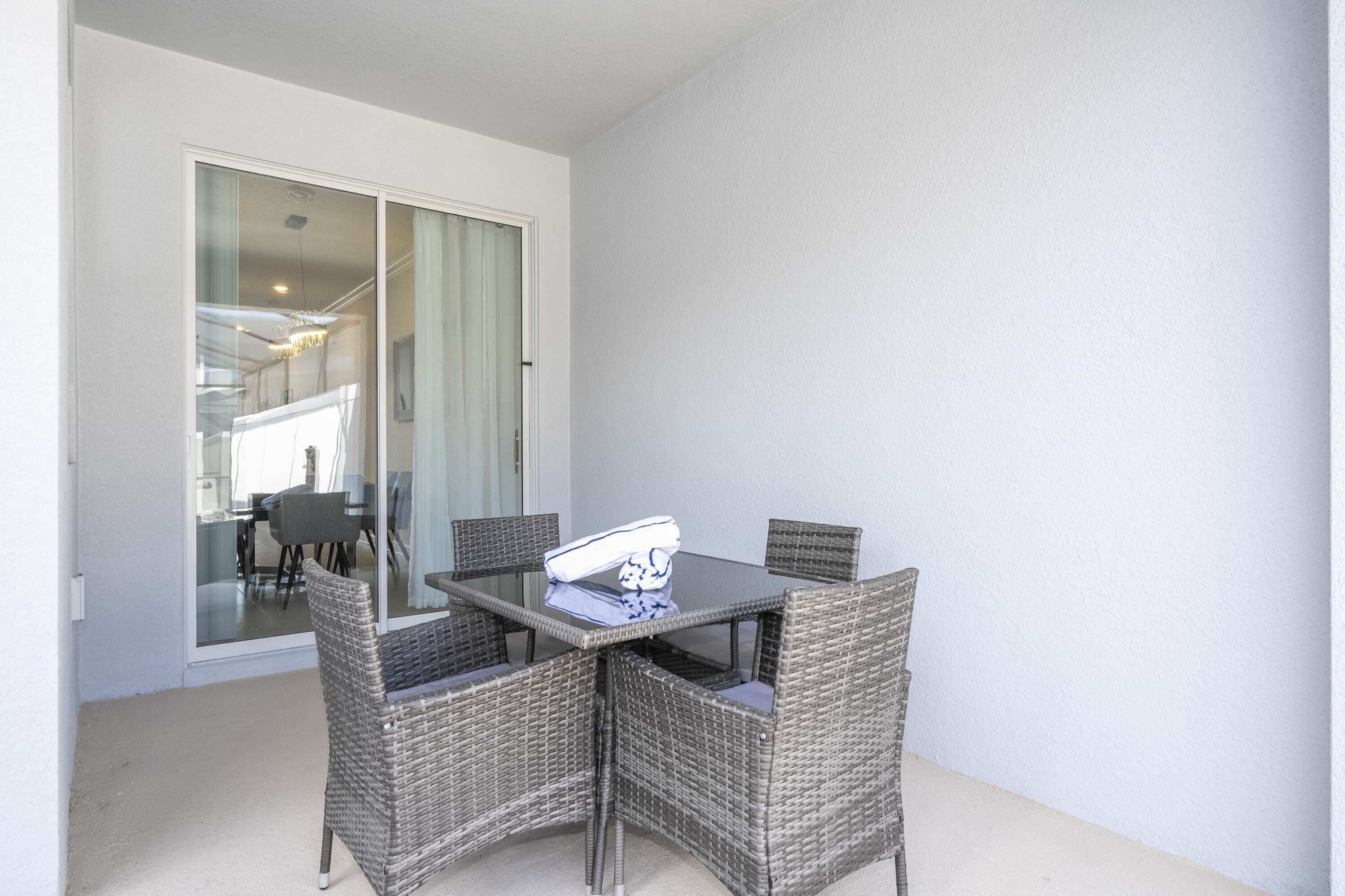 A dining area with a glass table and wicker chairs, a sliding glass door leading to a balcony, and a light-colored wall.
