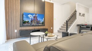 A modern living room with a flat-screen TV mounted on a wooden wall, a staircase with a 'PLAY' sign, and a pool table.