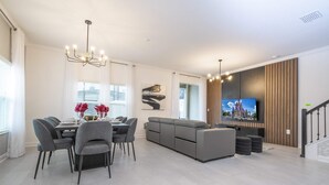 A modern living room with a dining area, grey sofas, a chandelier, and a flat-screen TV.