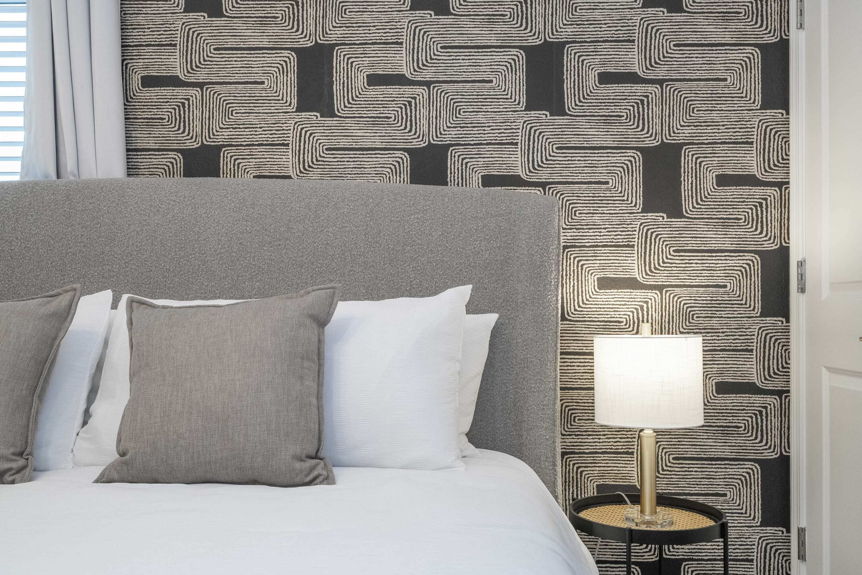 A bed with grey and white pillows, a black and white patterned wallpaper, and a small table with a lamp.