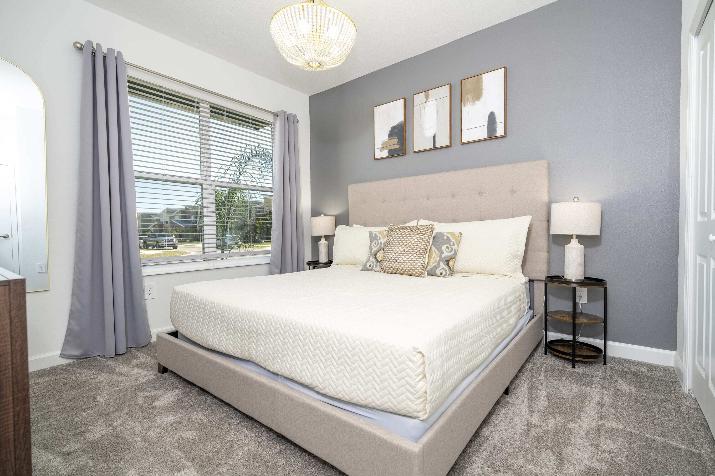 A bedroom with a large bed, two bedside tables with lamps, a chandelier, and a window with curtains.